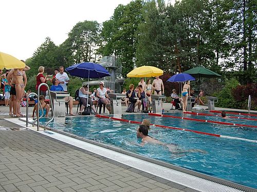 Am Rande eines Frei-Schwimmbeckens: Im Wasser schwimmen Leute. An den Startblöcken am Rande stehen und sitzen Menschen unter blauen, gelben und grünen Sonnenschirmen. Im Hintergrund ist eine spiralförmige große Rutsche zu erkennen. Bäume bilden den Hintergrund für die Szenerie.