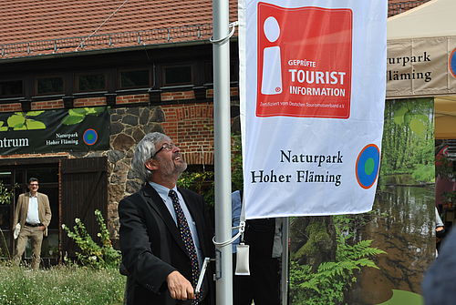 Ein Mann steht an einem Fahnenmasten und hisst eine Fahne mit der Aufschrift Information Naturpark Hoher Fläming. Im Hintergrund ein Fachwerkhaus mit Backstein.