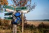 Eine Holzstele mit mit vielen Wanderwegehinweisen und behängt mit einem Wanderrucksack. Im Hintergrund sind Acker und Himmel zu sehen.