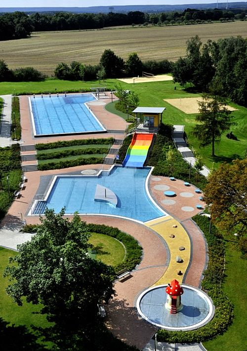 Schwimmbad mit Schwimm- und Planschbecken, Liegewiese und regenbogenfarbener breiter Rutsche aus der Vogelperspektive