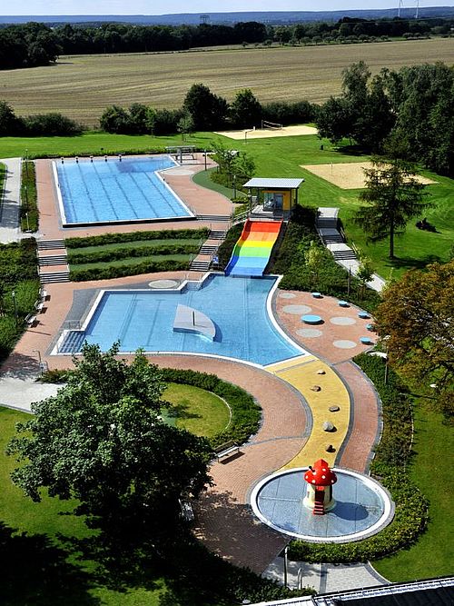 Schwimmbad mit Schwimm- und Planschbecken, Liegewiese und regenbogenfarbener breiter Rutsche aus der Vogelperspektive
