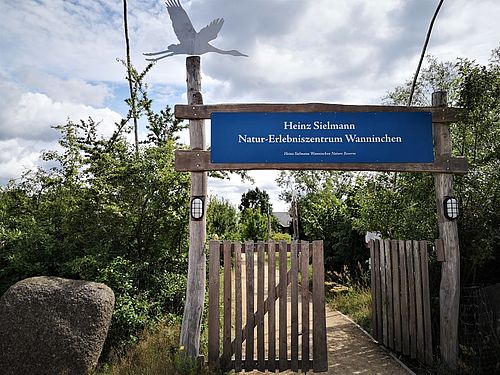 Das Tor zum Natur-Erlebniszentrum mit dem Namenszug. Auf einem Pfosten schwebt ein Kranich aus Metall.