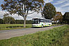 Ein Bus für den Personenverkehr fährt auf einer Landstraße, rechts eine Grasfläche mit Laub, links stehen Bäume.