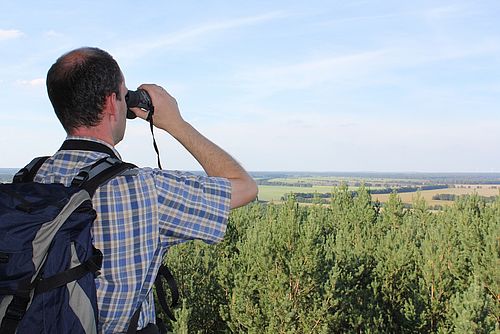 Ein Mann mit Rücksack hält ein Fernglas vor die Augen und blickt in die hügelige Landschaft mit viel Wald.