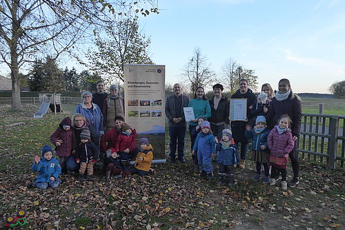 Eine Gruppe von Kindern und Erwachsenen haben sich für ein Gruppenfoto auf dem Kitagelände zusammengestellt.