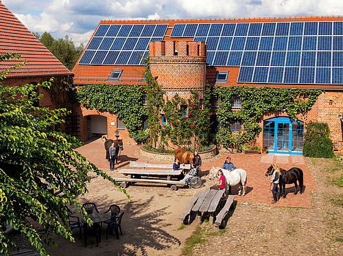Blick von oben auf einen Innenhof, dort stehen Menschen mit Pferden und Sitzgelegenheiten aus Holz umgeben von roten Klinkergebäuden und Solarmodulen auf dem Dach.