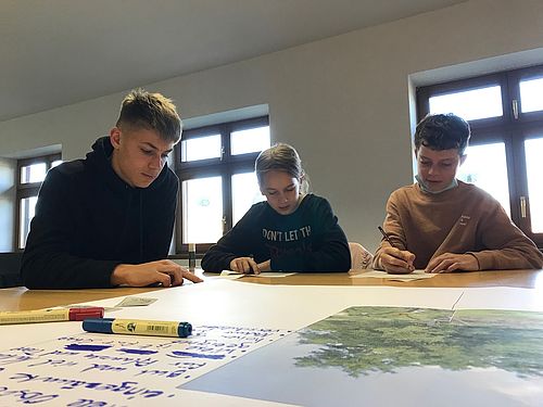 Drei Jugendliche schreiben etwas, auf dem Tisch liegt auch eine Karte.