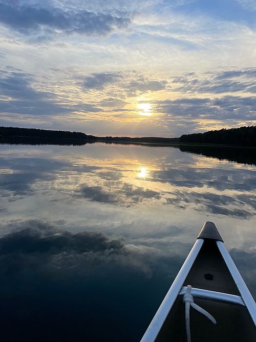 Man blick vom Kanadier aus auf den See in dem sich der beginnende Sonneuntergang spiegelt. Es ist eine weite Seefläche zu sehen und die Sonne ist noch relativ weit über der Erdoberfläche. Im Hintergrund sieht man den Waldrand. 