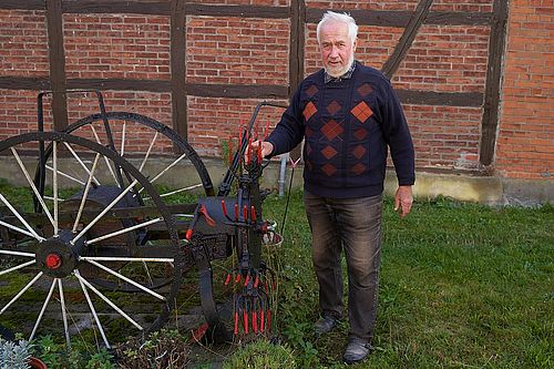 Ein älterer Herr steht neben einer alten Maschine, bestehend aus zwei großen Rädern und einigen Gabeln, die die Kartoffeln aufsammeln.