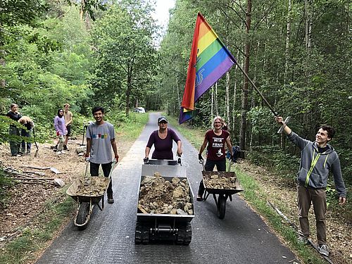 Drei junge Leute mit Schubkarre bzw. kleinem Dumper voller grober Steine. 