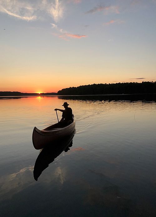 Eine Person sitzt in einem Kanadier und fährt auf einem See, im Hintergrund der Sonnenuntergang und die Konturen eines Waldgebietes am Ufer des Sees. 