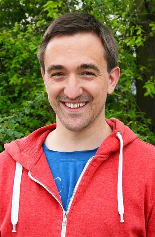 Porträt eines jungen lächelnden Mannes in roter Sweatjacke und blauem T-Shirt. Im Hintergrund Grün.