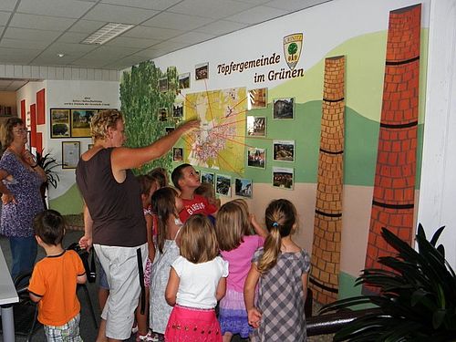 eine Frau zeigt einer Gruppe Kinder etwas an einer mit Karte, Bildern und Zeichnungen gestalteten Wand