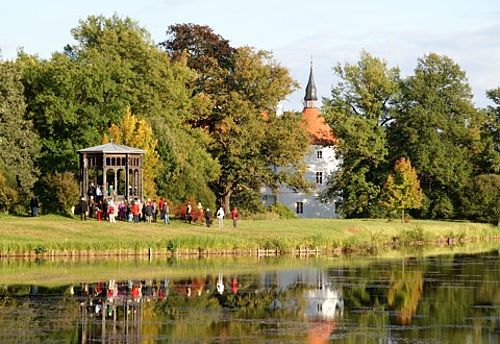 Parklandschaft: Ein Pavillon mit einer Gruppe Menschen und das zwischen alten Bäumen hervorlugende Schloss spiegeln sich im Teich