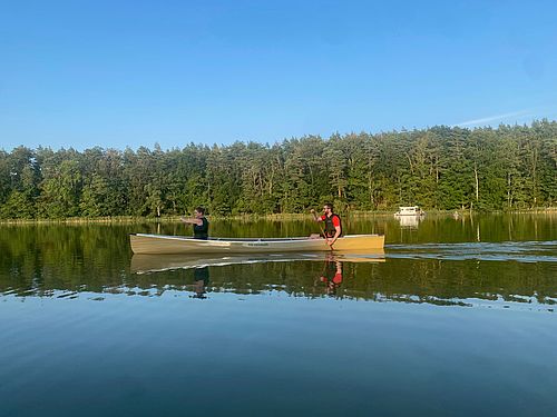 Zwei Personen in einem Tandem-Kanadier auf einem See, im Hintergrund sieht man einen Wald. Der blaue, wolkenlose Himmel spiegelt sich im Wasser unter dem Kanadier. 