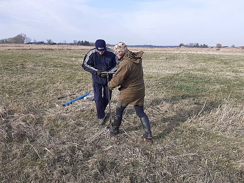 Zwei Menschen auf einer trockene Wiese bohren ein Loch in den Boden.
