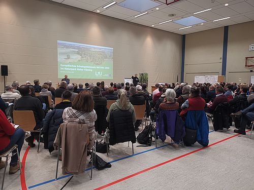 Viele Menschen sitzen in einer großen Halle und schauen auf eine Leinwand.