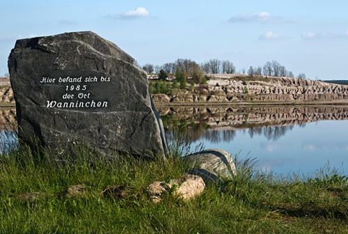 Hinter dem Gedenkstein mit der Aufschrift "Hier befand sich bis 1985 der Ort Wanninchen" spiegeln sich im Wasser des Schlabendorfer Sees der blaue Himmel und die zerklüfteten Böschungen des gegenüberliegenden Ufers.