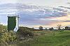 Ehemaliger Grenzturm bei Gandow inmitten einer weiten, grünen Landschaft bei Sonnenuntergang. Der Turm ist teilweise mit Efeu bewachsen und steht auf einem Deich, umgeben von Wiesen, Sträuchern und vereinzelten Bäumen.
