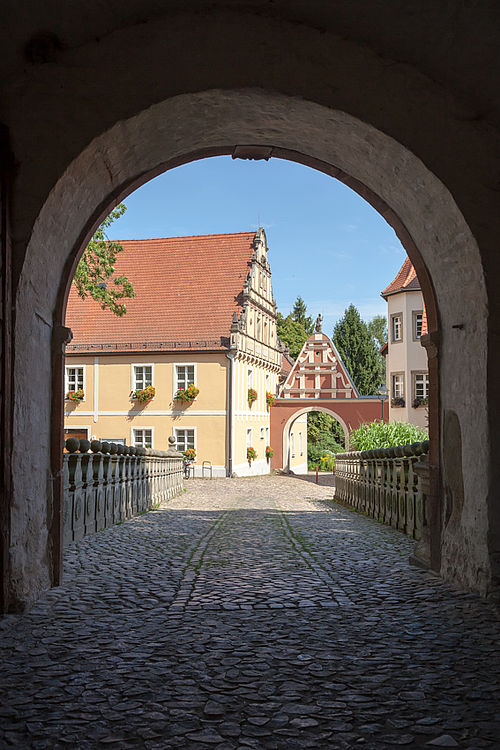 Blick aus einer gewölbten Tordurchfahrt auf ein mit Blumenkästen geschmücktes Gebäude und ein kleines Tor in einer Siedlung.
