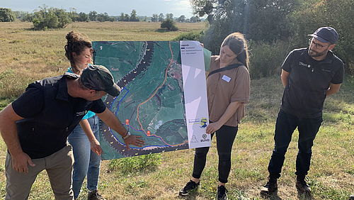 Drei Personen stehen auf einer Wiese vor einer großformatigen Luftbildkarte; Maria Lindow hält die Karte, Jan Schormann steht rechts daneben