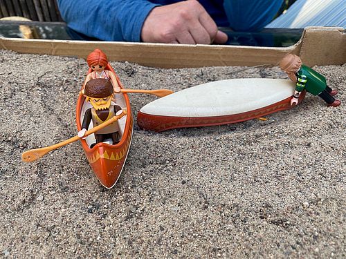 Drei Playmobilfiguren im Sandkasten, eine schiebt ein Playmobil-Boot, zwei sitzen gemeinsam in einem Playmobil-Kanadier. 